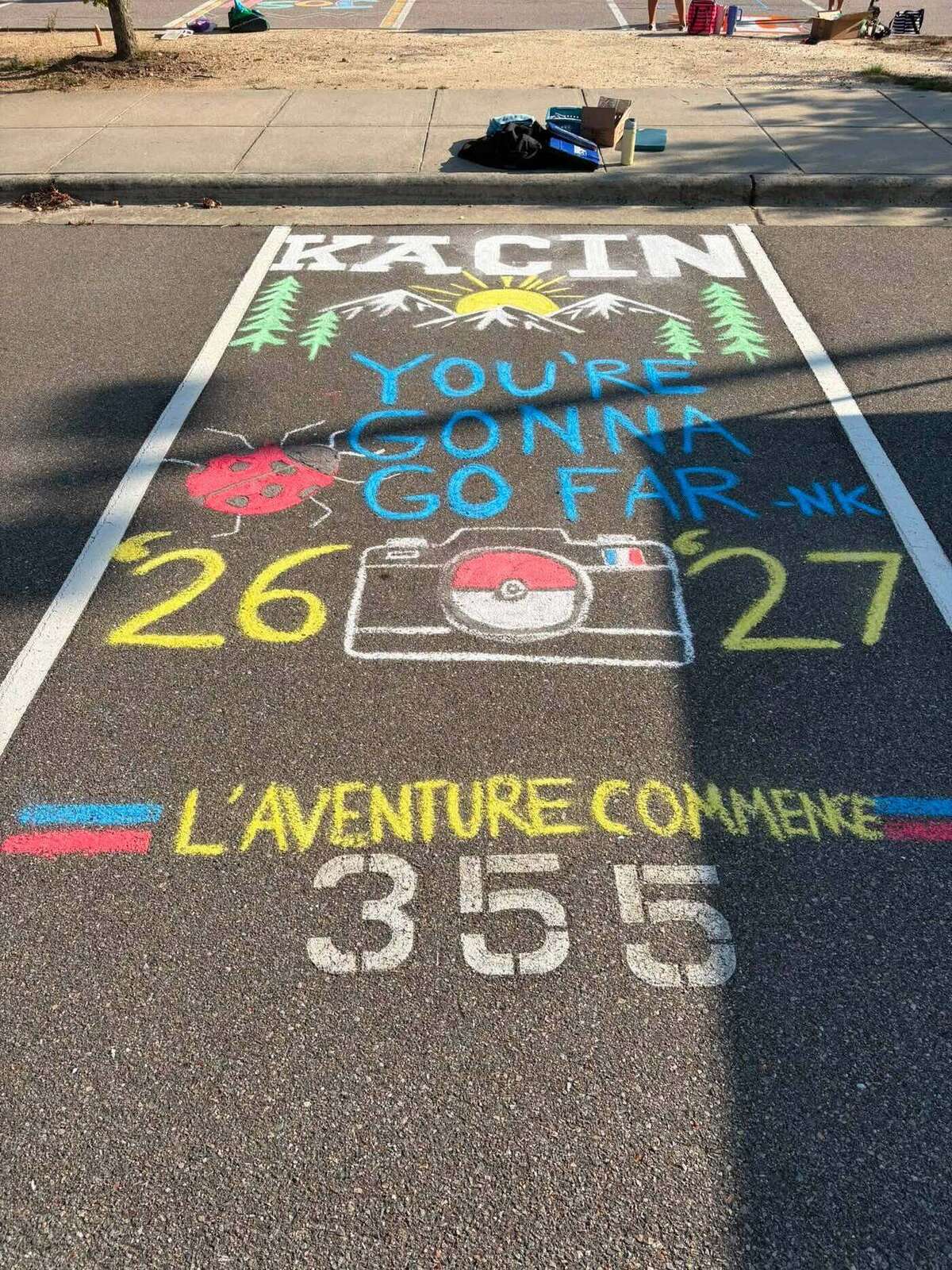 A chalked parking space reading You're Gonna Go Far, with mountains, a sunrise, pine trees and a camera, above the words L'aventure Commence
