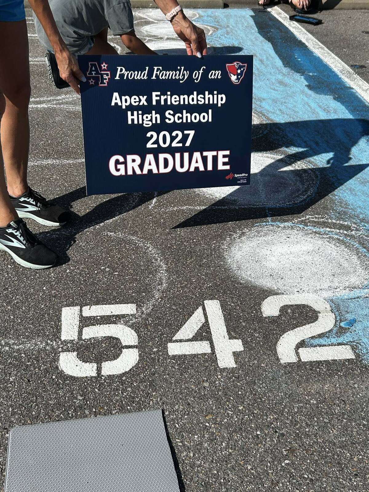 A Proud Family of an Apex Friendship High School 2027 Graduate yard sign standing over a freshly chalked parking space